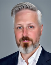 A man with gray hair and beard in a navy blue suit jacket and white shirt stands against a gray background, appearing ready for discussions on roadway design with state and local authorities.