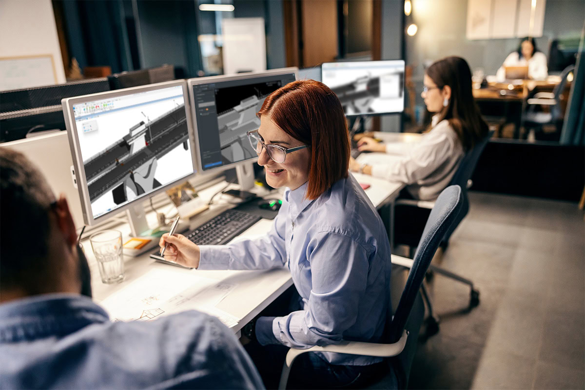 Three people working at desks with computer monitors displaying 3D models in MicroStation. A woman with red hair wearing glasses is in focus, smiling and talking to a colleague. An office environment is visible, reflecting the collaborative spirit of users as they work on projects for 2024.