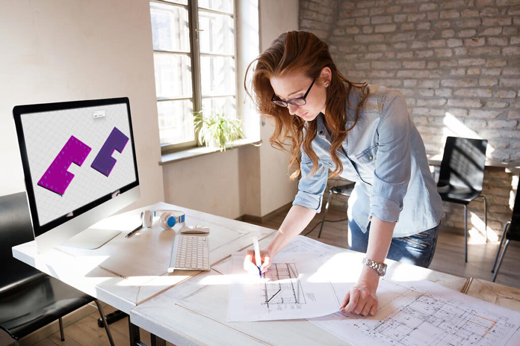 A woman with glasses reviews architectural plans on a desk while a computer screen displays a 3D model of a building in MicroStation. She works in a well-lit room with brick walls, showcasing the latest in design for 2024.